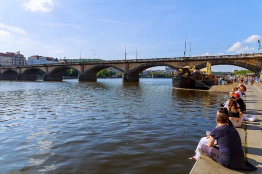 22 May 2022 Prague, Czech Republic. Embankment near river is popular meeting place for locals and tourists. People watch ships and enjoy view of river and the bridge.