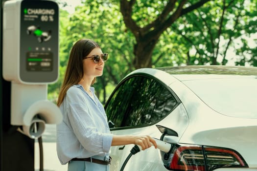 Young woman recharging battery for electric car during road trip travel EV car in natural forest or national park. Eco friendly travel during vacation and holiday. Exalt