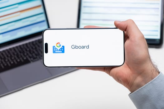 Gboard application logo on the screen of smart phone in mans hand, laptop and tablet are on the table in the background, December 2023, Prague, Czech Republic.