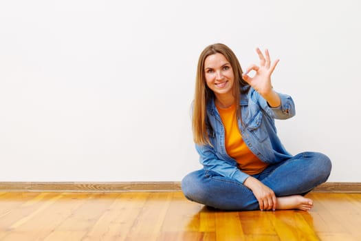 Smiling woman in jeans and denim jacket sitting on the floor giving an OK hand sign, expressing satisfaction.