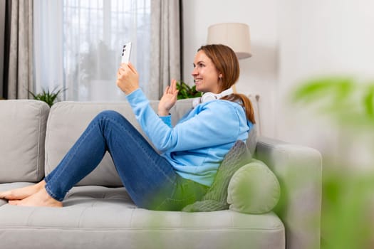 Relaxed woman lying on couch at home, happily video calling with her smartphone