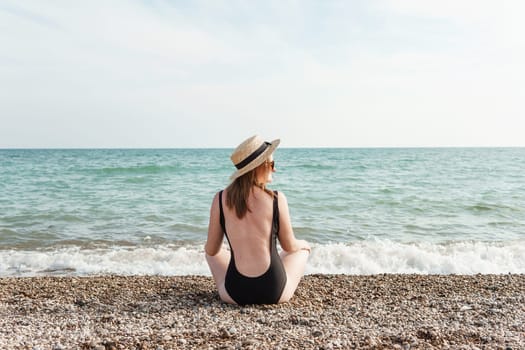 Woman in black swimsuit and straw hat on beach, seaside. View from the back. Summer vacation at the sea