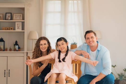 Happy family portrait with lovely little girl smiling and looking at camera, lovely and cheerful parent and their daughter sitting together in living room at home with warm daylight. Synchronos