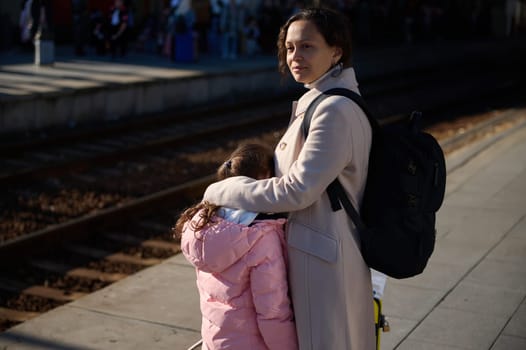 Young woman with suitcase and backpack, mother and her daughter in warm coats, standing by railway in train station, waiting for train. Railroad transport concept. People. Travel and trip. Lifestyle