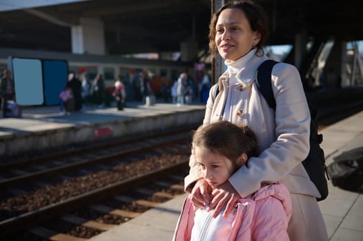Young multiethnic woman, a mother and her little daughter waiting for the train, standing on the platform at the railway station.