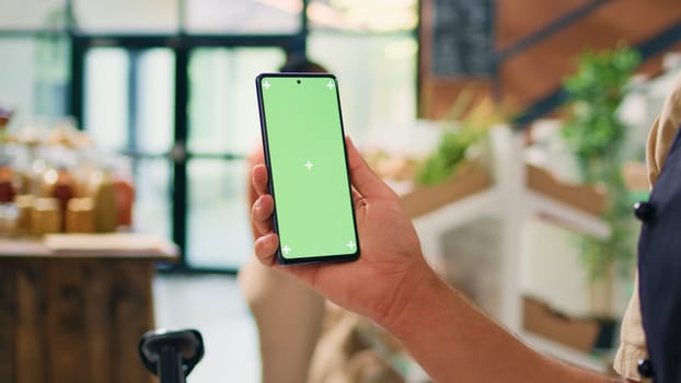 Owner uses greenscreen on smartphone, showing isolated chromakey template on device in local organic zero waste eco market. Young vendor presents copyspace mockup display.