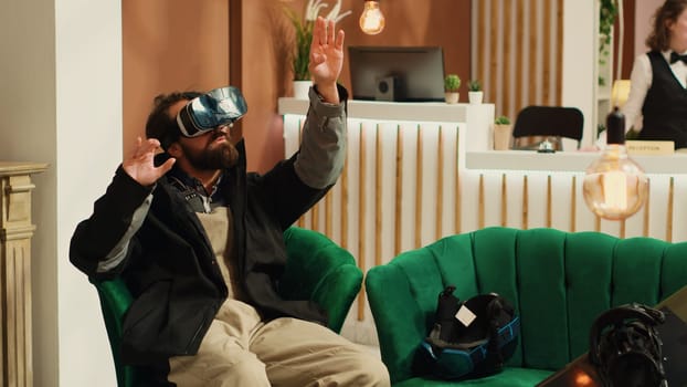 Traveler uses virtual reality technology in lounge area, having fun with vr interactive glasses while he wears winter jacket. Person having helmet and snowboard next to him, prepare to go skiing.