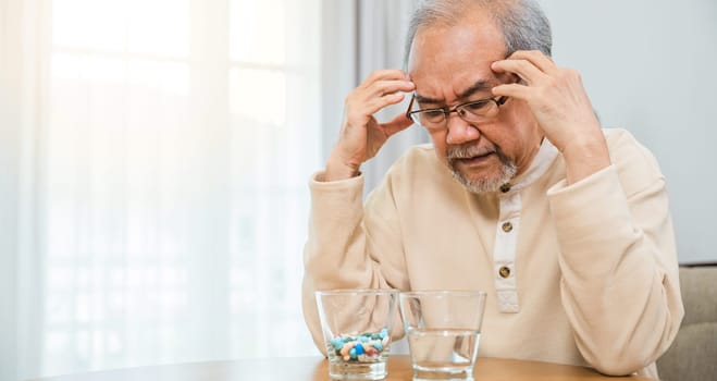 Asian senior old man grey haired sad sitting in chair hand hold head with many medicines and pills for taking, elderly man sick have headache looking pill glass on at home, Elderly healthcare