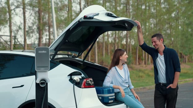 Couple recharge their EV car with electric battery charging station at rest stop during their road trip travel during autumnal season to national park. Relaxing holiday with eco friendly car. Exalt