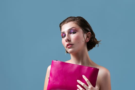 Portrait of topless young female model with short brown hair and eyeshadow holding pink purse while looking at camera over blue background