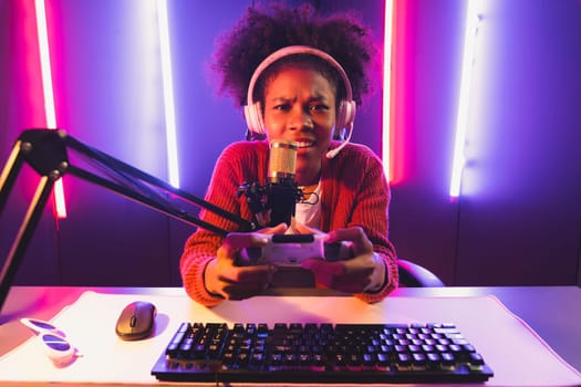 Host channel of gaming streamer, African girl playing online game with joystick, talking with viewers media online on microphone. Esport skilled team players in neon color lighting room. Tastemaker.