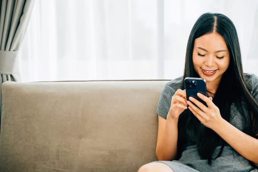 Asian woman relaxes on a cozy couch texting and shopping online via smartphone. Engaged in communication leisure and digital activities from home.