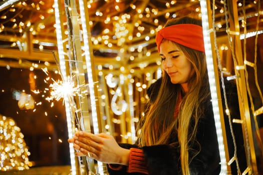 Woman holding sparkler night while celebrating Christmas outside. Dressed in a fur coat and a red headband. Blurred christmas decorations in the background. Selective focus.