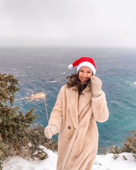 Outdoor winter portrait of happy smiling woman, light faux fur coat holding heart sparkler, posing against sea and snow background.