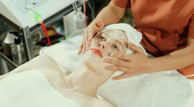 Portrait of beautiful caucasian woman having facial massage with homemade facial mask while lies on spa bed surrounded by beauty electrical equipment and peaceful nature environment. Tranquility.