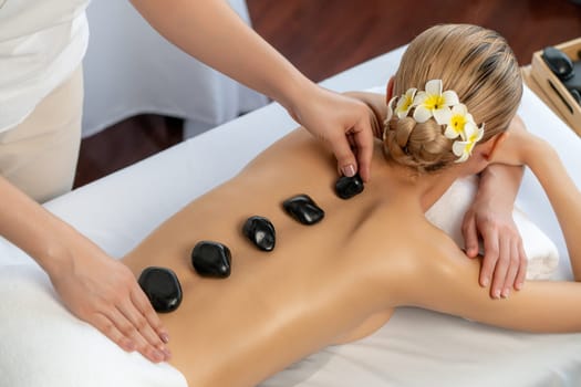 Hot stone massage at spa salon in luxury resort with day light serenity ambient, blissful woman customer enjoying spa basalt stone massage glide over body with soothing warmth. Quiescent