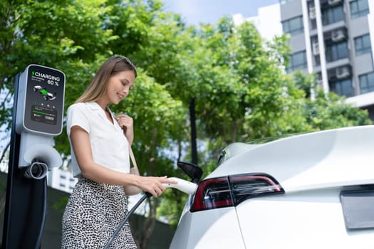 Young woman travel with EV electric car charging in green sustainable city outdoor garden in summer. Urban sustainability lifestyle by green clean rechargeable energy of electric BEV vehicle innards