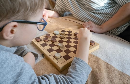 Close-up of a caring middle-aged grandmother playing checkers with a child, a boy, while lying on the bed. Happy family enjoying an interesting wooden board game at home
