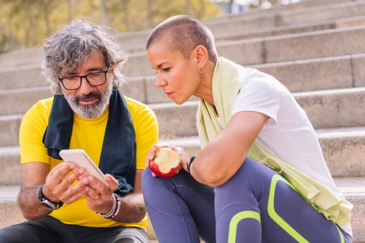 senior sporty man showing mobile phone to his personal trainer who is eating an apple, concept of technology and healthy lifestyle