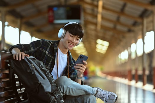 Happy young man in headphone using mobile phone during waiting for train at railway station.