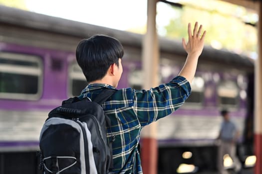 Happy young Asian man waving hand to friend while at a railroad station platform.