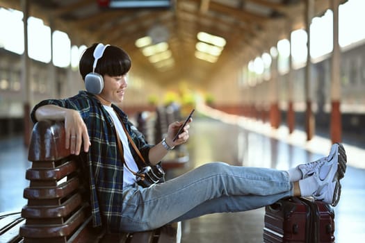 Happy young man in headphone using mobile phone during waiting for train at railway station.