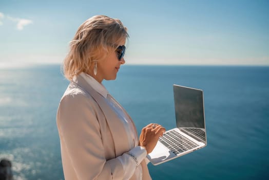 Freelance women sea working on the computer. Good looking middle aged woman typing on a laptop keyboard outdoors with a beautiful sea view. The concept of remote work