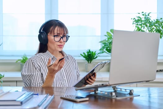 Middle-aged woman in headphones working at computer in home office, looking at web cam talking. Online meeting, remote work teaching financial legal advice interview blogging freelance, mental therapy