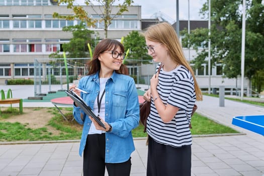 Talking female teacher and teenage high schoolgirl outdoor, school building background. Meeting communication student girl with backpack and mentor counselor. Education, adolescence, learning concept