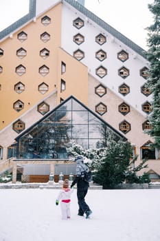 Mother with a small child walks by the hand past a triangular hotel in the snow. Back view. High quality photo