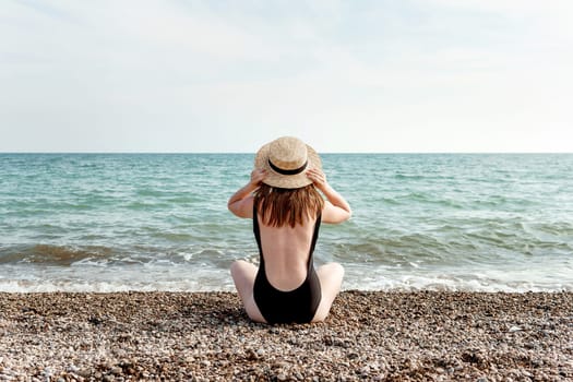 Woman in black swimsuit and straw hat on beach, seaside. View from the back. Summer vacation at the sea