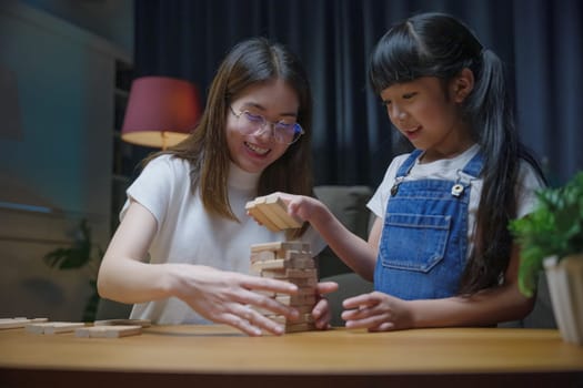 Smiling woman help teach child play build constructor of wooden blocks, Asian young mother playing game in wood block with her little daughter in home living room at night time before going to bed