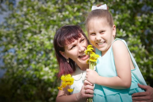 Happy mother and daughter enjoying rest, playing and fun on nature on a green lawn with dandelions and blooming apple tree on background. Woman and girl resting outdoors in summer and spring day