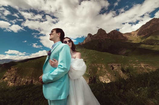 wedding couple walks in the mountains. the bride hugs tenderly with her eyes closed the groom from the back against the backdrop of green rocky mountains and blue sky, close-up.