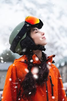 Smiling young woman, portrait, standing and enjoying snowfall, wearing protective clothing, helmet and goggles. Winter sports resort.