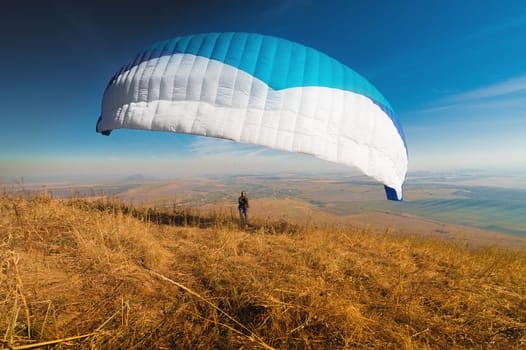 A paraglider takes off from a mountainside with a blue and white canopy and the sun behind. A paraglider is a silhouette. The glider is sharp, with little wing movement. A male paraglider launches a paraglider into the air.