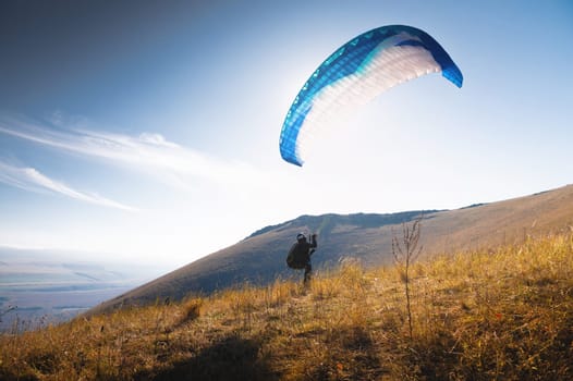 A paraglider takes off from a mountainside with a blue and white canopy and the sun behind. A paraglider is a silhouette. The glider is sharp, with little wing movement. A male paraglider launches a paraglider into the air.