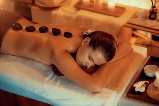 Hot stone massage at spa salon in luxury resort with warm candle light, blissful woman customer enjoying spa basalt stone massage glide over body with soothing warmth. Quiescent