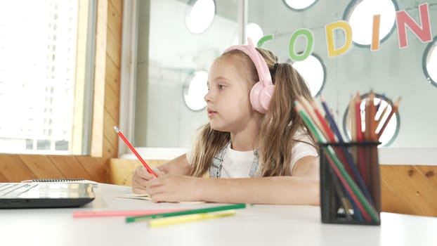 Cute smart girl doing classwork or online learning while listen to teacher. Happy student writing, drawing, working on paper while wearing headphone with laptop and colorful color pencils. Erudition.