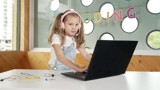 Little young cute girl using laptop to working or playing games. Student working on computer while looking and smiling at camera. Caucasian child attend online class by using headphone. Erudition.