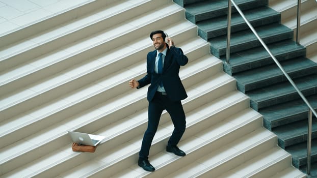 Professional business man standing at stair while moving to music from headphone. Skilled project manager dancing while listening relax sound with laptop placed on stairs. Modern dance. Exultant.