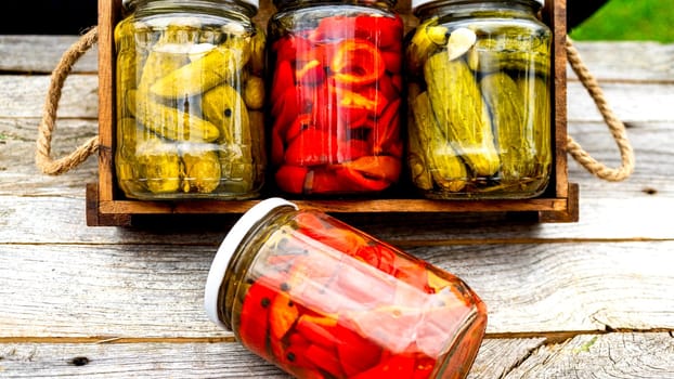 Glass jars with pickled red bell peppers and pickled cucumbers (pickles) isolated in wooden crate. Jars with variety of pickled vegetables. Preserved food concept in a rustic composition.