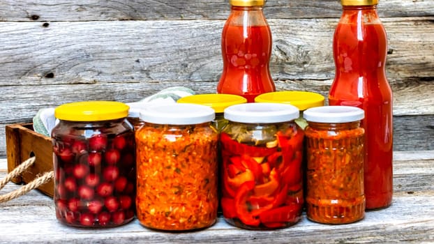 Jars with variety of canned vegetables and fruits, jars with zacusca and bottles with tomatoes sauce. Preserved food concept in a rustic composition.