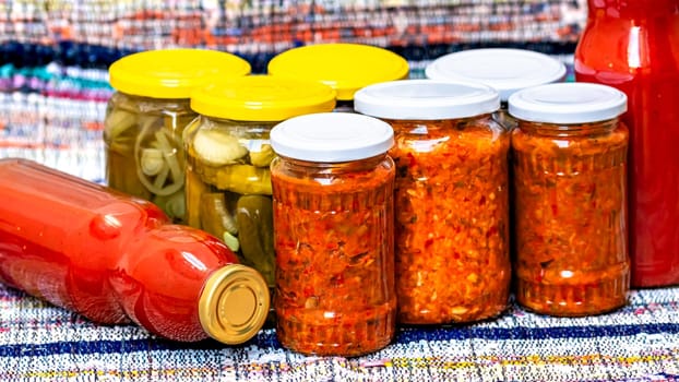 Glass jars with pickled red bell peppers and pickled cucumbers (pickles) isolated. Jars with variety of pickled vegetables, jars with zacusca and bottles with tomatoes sauce. Preserved food concept.