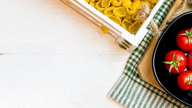 Top view of pasta in a wooden crate and fresh ripe cherry tomatoes in a black bowl on a rustic white wooden table. Ingredients and food concept