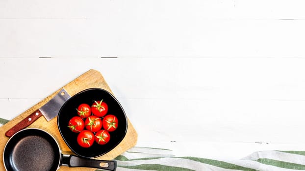 Top view of knife, small pan and fresh ripe cherry tomatoes in small black bowl on a rustic white wooden table. Ingredients and food concept