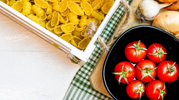 Top view of onions, garlic, pasta in a wooden crate and fresh ripe cherry tomatoes on a rustic white wooden table. Ingredients and food concept