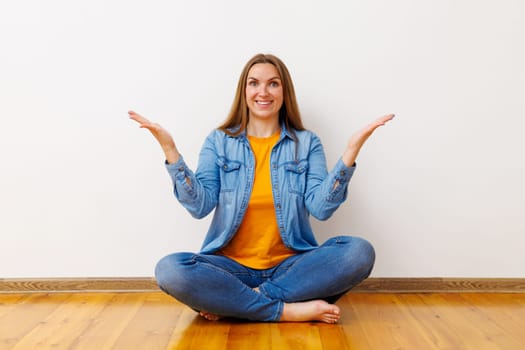 A young woman sitting cross-legged on a wooden floor, looking indecisive with hands raised in a balancing gesture.
