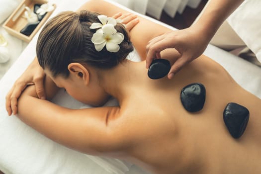 Hot stone massage at spa salon in luxury resort with day light serenity ambient, blissful woman customer enjoying spa basalt stone massage glide over body with soothing warmth. Quiescent