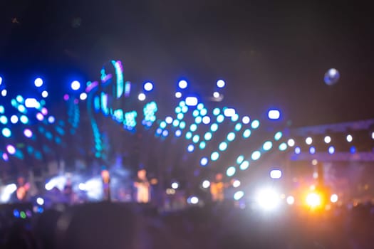 The night is alive with music and excitement at a concert festival main event. A cheering unrecognizable crowd gathers in front of the brightly lit stage and the lens flare adds to lively atmosphere.